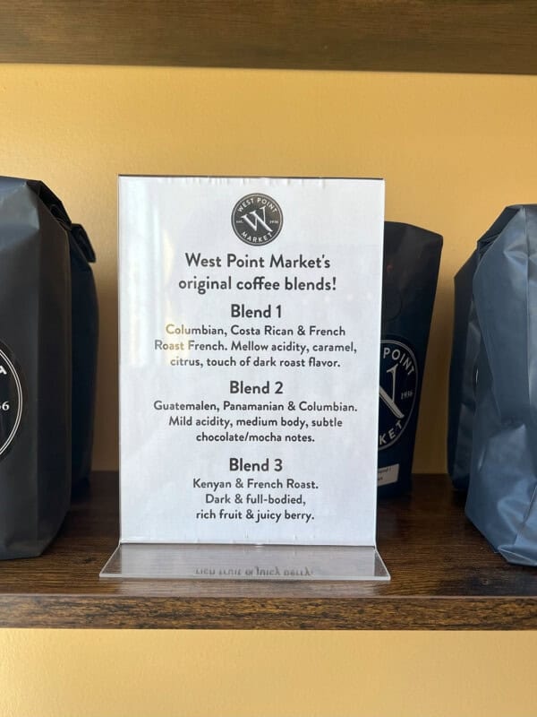 A wooden shelf features a sign describing three WPM Original Coffee Blends, detailing each blend’s origin, flavor notes, and roast profile, with coffee bags displayed on either side of the sign.