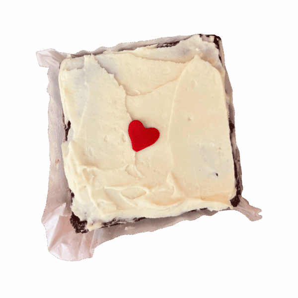 A WPM Original Triple Layer Brownie with white frosting and a red heart-shaped decoration sits on parchment paper against a white background.