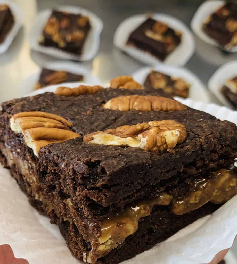 A close-up of a rich chocolate brownie filled with caramel and pecans, topped with whole pecans. Several other brownies on paper liners are blurred in the background.