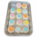 A metal baking tray holds fifteen Thumbprints cookies, each topped with pastel icing in pink, orange, blue, or yellow. The cookies are arranged in three rows of five.