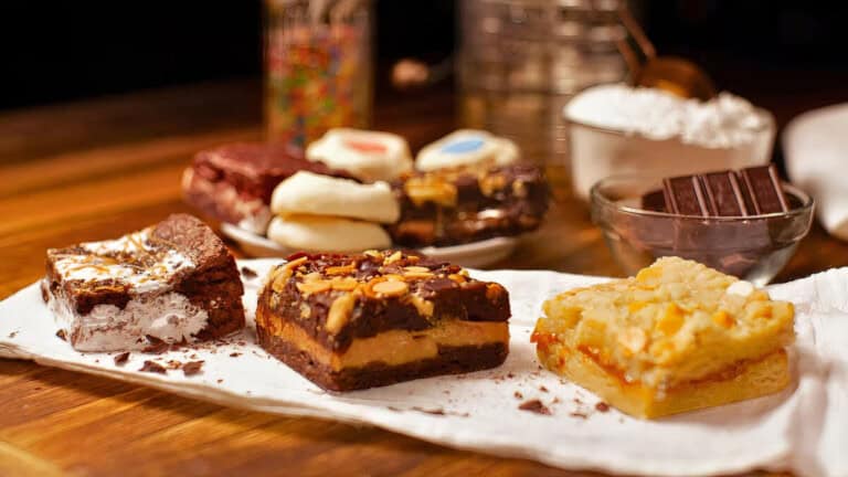 A close-up of assorted dessert bars and cookies arranged on a white napkin, with ingredients like chocolate, flour, and sprinkles blurred in the background on a wooden surface.