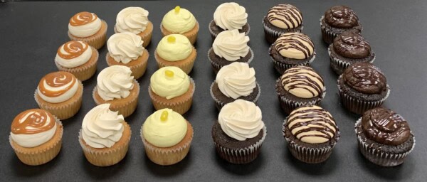 Pandora's Cupcakes - Daily 6 Flavors: Five rows of four cupcakes each, topped with caramel drizzle, lemon candy, chocolate drizzle, and plain swirls, beautifully displayed on a black surface.