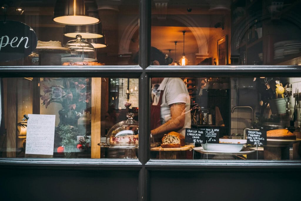 A person stands behind a bakery window arranging pastries and cakes on display, with handwritten signs and a note visible beside glass domes holding desserts. Warm lights illuminate the cozy interior.