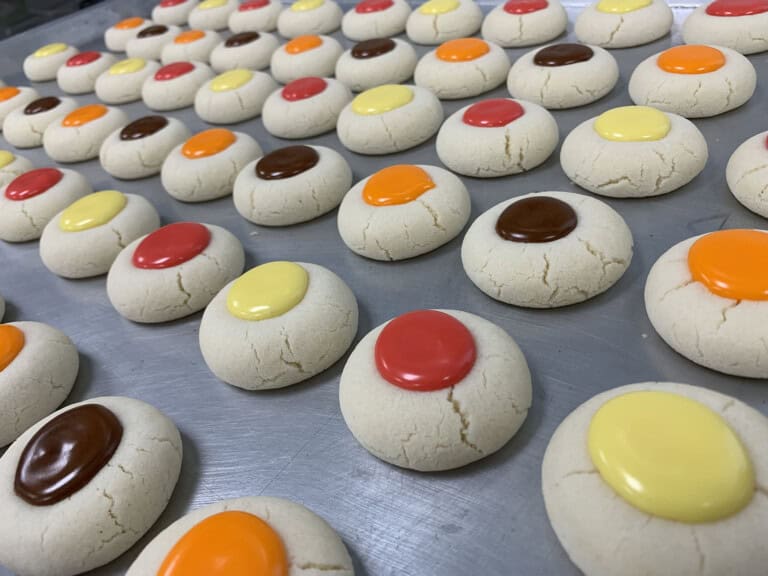Rows of round, light-colored sugar cookies topped with colorful, glossy candy buttons in red, yellow, orange, and brown are arranged neatly on a metal baking sheet.
