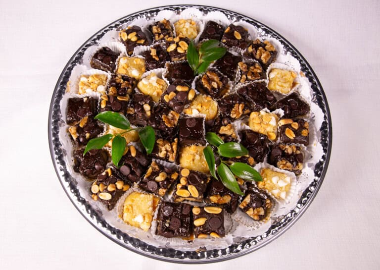 A round silver tray filled with assorted chocolate and nut squares, each placed in a white paper cup, garnished with a few green leaves, on a white tablecloth.