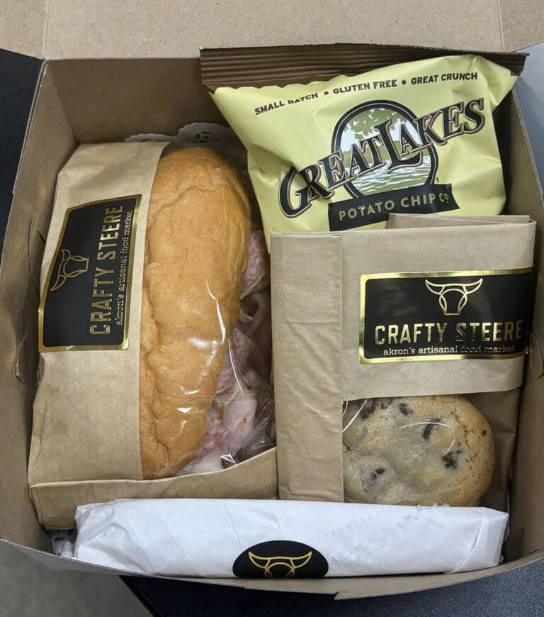 A boxed lunch containing a sandwich, a bag of Great Lakes potato chips, a chocolate chip cookie, a wrapped utensil, and items labeled “Crafty Steere” on brown paper packaging.