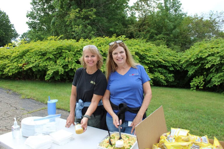 Two women smiling while serving food at an outdoor table. Plates, utensils, and snacks are on the table. Green bushes and trees are in the background. Both women are dressed casually and appear to be enjoying the event.