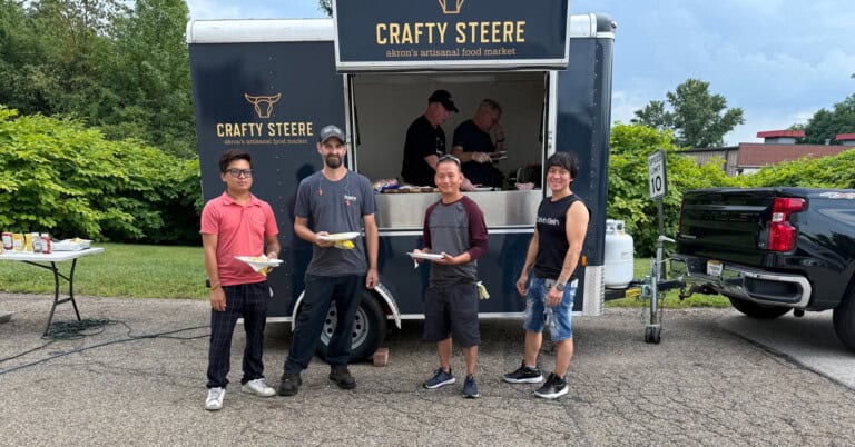 Four people stand smiling in front of a Crafty Steere food truck, holding plates of food, with two workers visible inside the truck. They are outdoors on a paved area with greenery in the background.