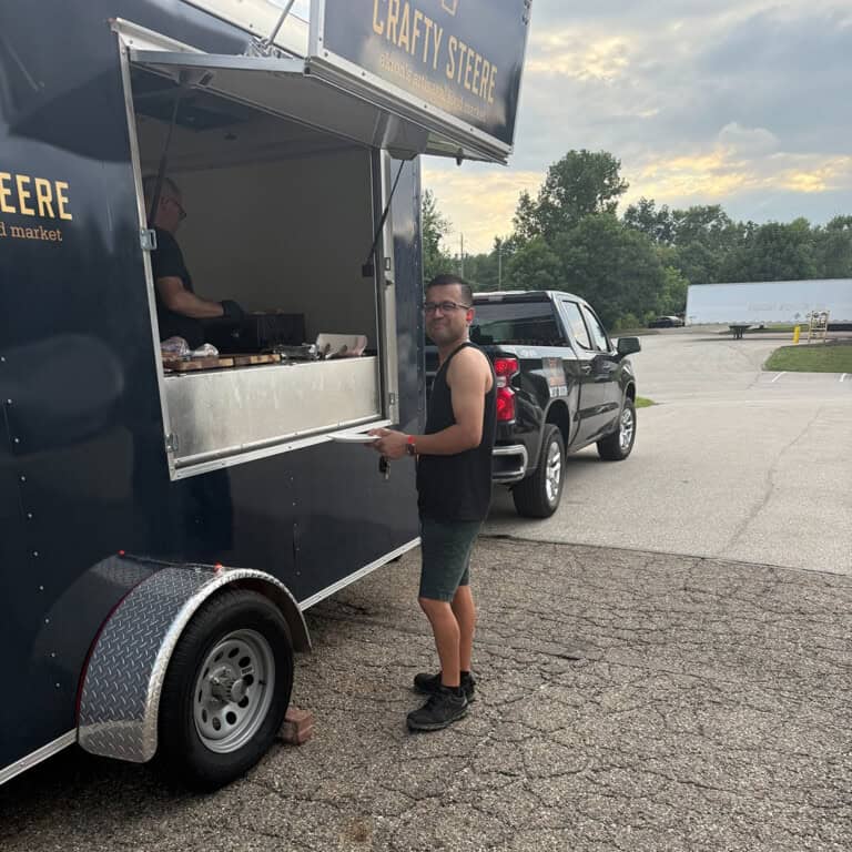 A man in casual clothing stands at the serving window of a dark food truck parked on a paved lot, holding food. Another person is visible inside the truck, preparing or serving food. Trees and vehicles are in the background.