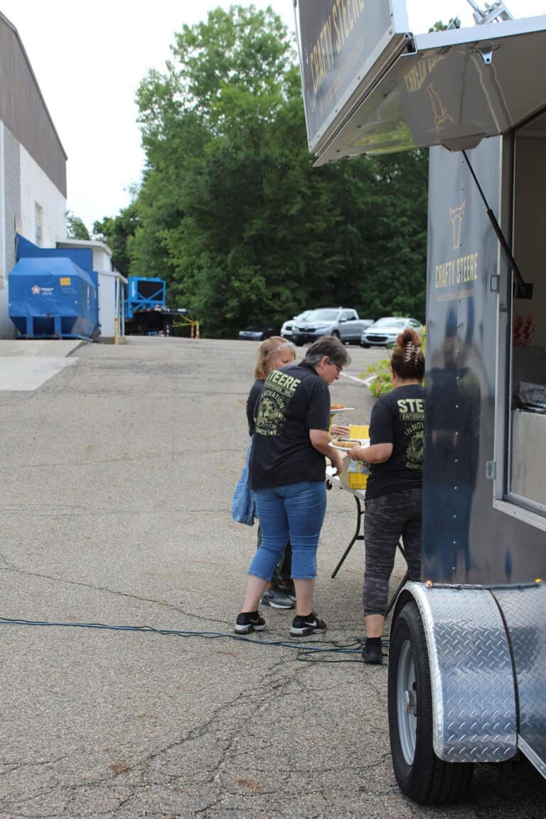Three people in matching black Crafty Steer shirts prepare food at an outdoor food truck. The scene takes place in a parking lot near a dumpster and several parked cars, with trees in the background.