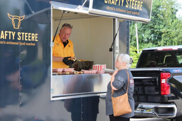 A man in a yellow shirt serves food from a blue food truck labeled Crafty Steere to a woman standing outside, holding a brown purse, with a pickup truck parked nearby.