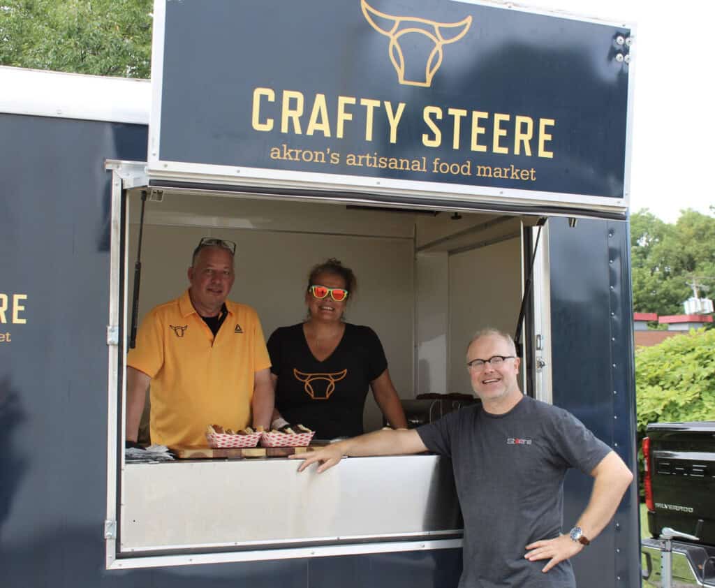 Three people smile at a food truck window labeled Crafty Steere, akron’s artisanal food market. Two stand inside the truck, while one stands outside, leaning on the counter. Food trays are displayed on the window ledge.