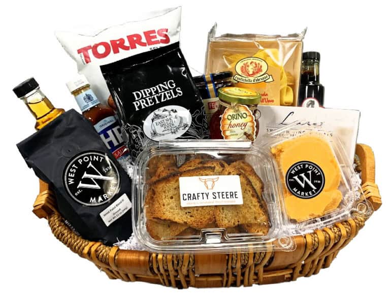 The Gift Basket - Small is a wicker basket filled with gourmet crackers, cheese, chips, pretzels, honey, sauces, and two bottles, all neatly arranged with labels visible.