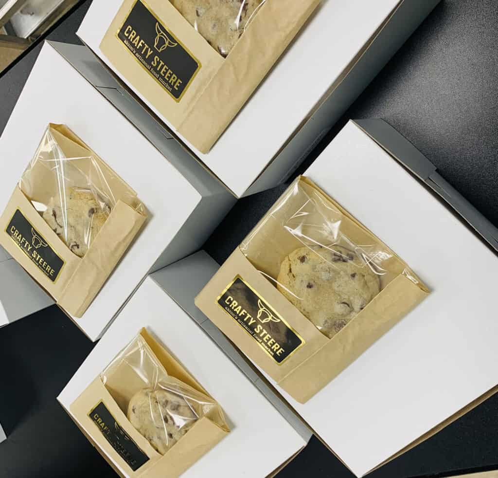 Several white boxes with see-through pockets, each containing a chocolate chip cookie in a clear bag with a “Crafty Steere” label, arranged diagonally on a black surface.