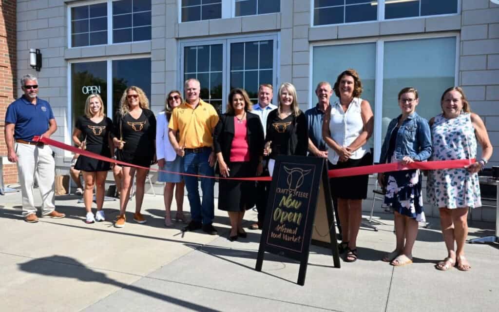 A group of people stand outside a storefront holding a red ribbon for a grand opening ceremony. A signboard in front reads Curts Steer New Open - Local Food Market. All are smiling at the camera.