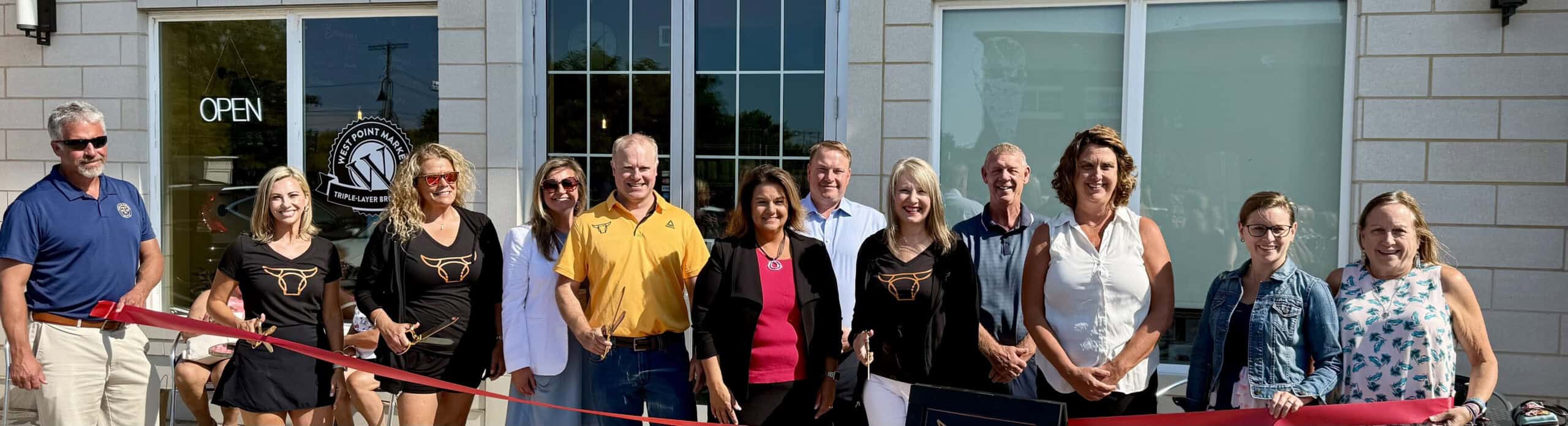 A group of people stand in front of a building for a ribbon-cutting ceremony, smiling at the camera. Several hold a large red ribbon, with sunlight shining on them. The building behind has large windows and a sign.