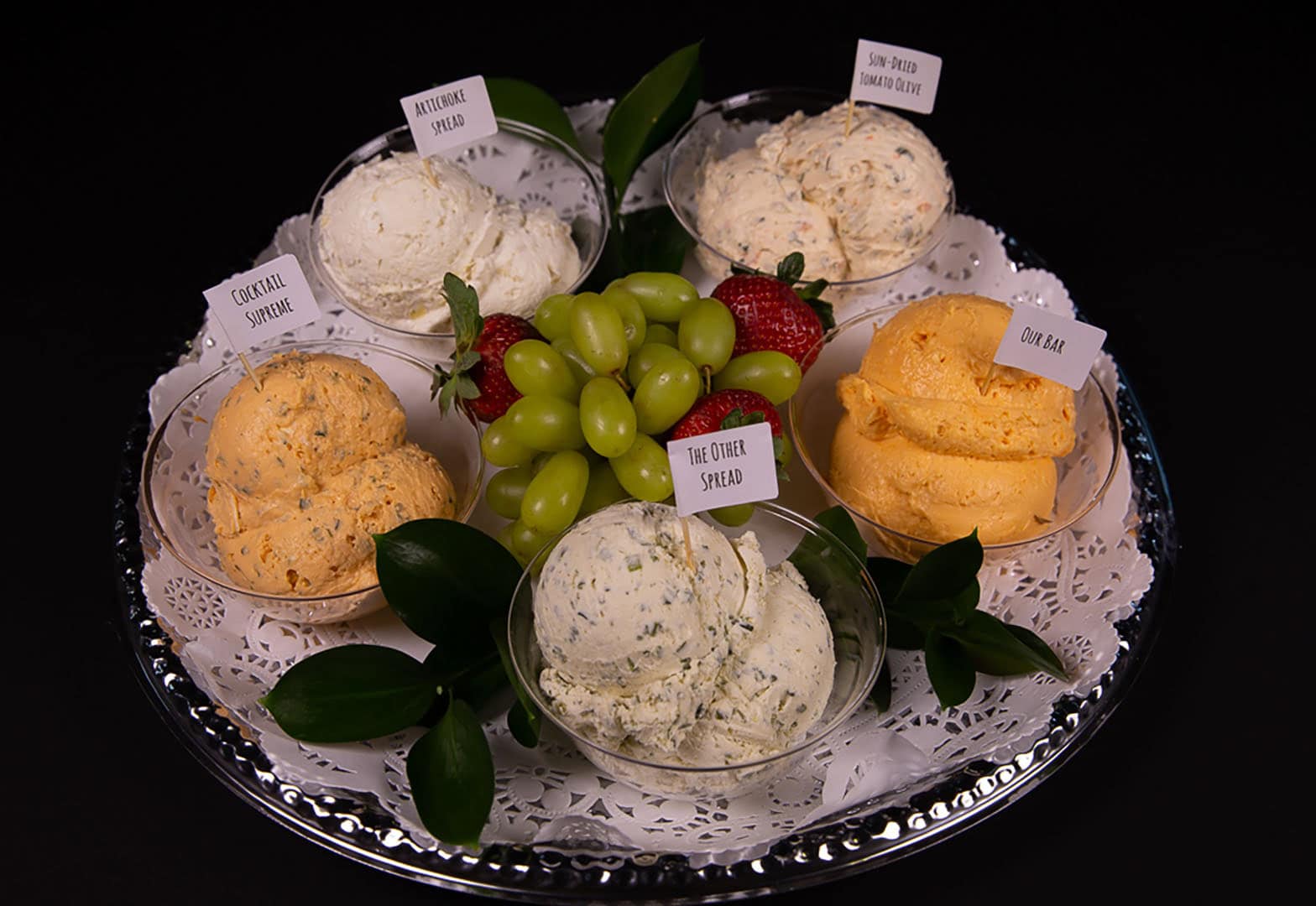 A silver tray with five bowls of assorted cheese spreads, each labeled, arranged around green grapes and strawberries, with green leaves as garnish on a decorative white doily.