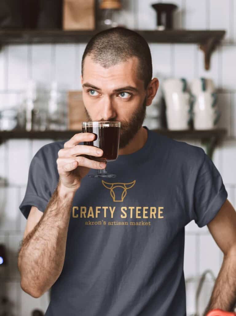 A man with short hair and a beard drinks coffee from a glass mug. He is wearing a blue Crafty Steere Akrons Artisan Market t-shirt and standing in a kitchen with shelves and mugs in the background.