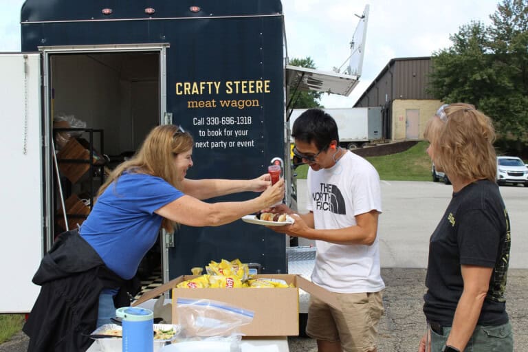 A woman enthusiastically squirts ketchup onto a mans food as he holds a plate, while another woman watches and smiles. They are standing outside near a food truck labeled Crafty Steere meat wagon.