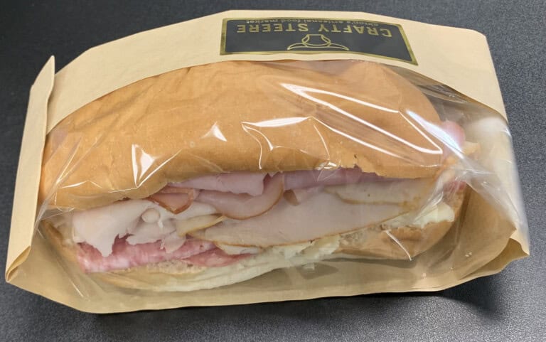 A packaged sandwich with sliced turkey and ham on a soft bun, wrapped in clear plastic and brown paper, labeled CRAFTY STREET. The sandwich is placed on a dark surface.