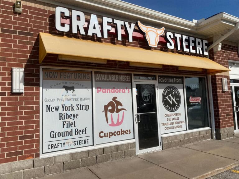 Storefront of Crafty Steere with large signs advertising New York strip, ribeye, filet, ground beef, Pandora’s Cupcakes, and cheese spreads. The building has a yellow awning and brick exterior.