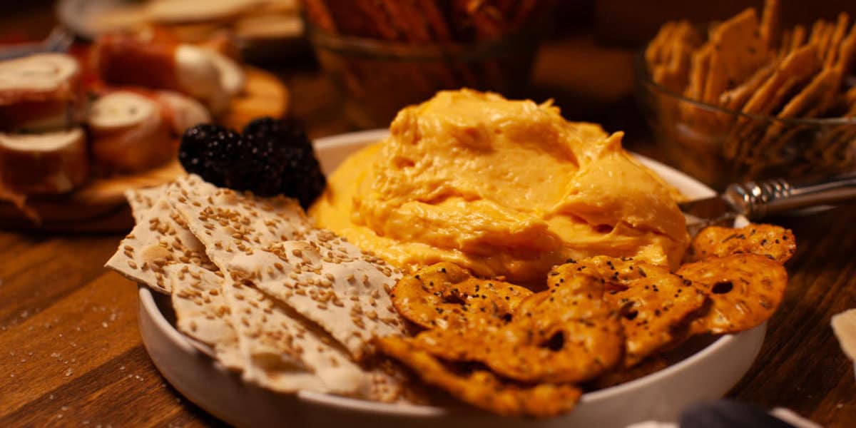 A white plate with a mound of cheese spread, surrounded by seeded flatbread crackers, seasoned pretzel chips, and blackberries, set on a wooden table with more snacks in the background.