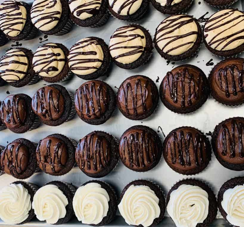 A tray of assorted cupcakes arranged in rows, with chocolate cupcakes topped with chocolate drizzle, vanilla cupcakes with white frosting swirls, and some cupcakes with beige frosting and chocolate drizzle.