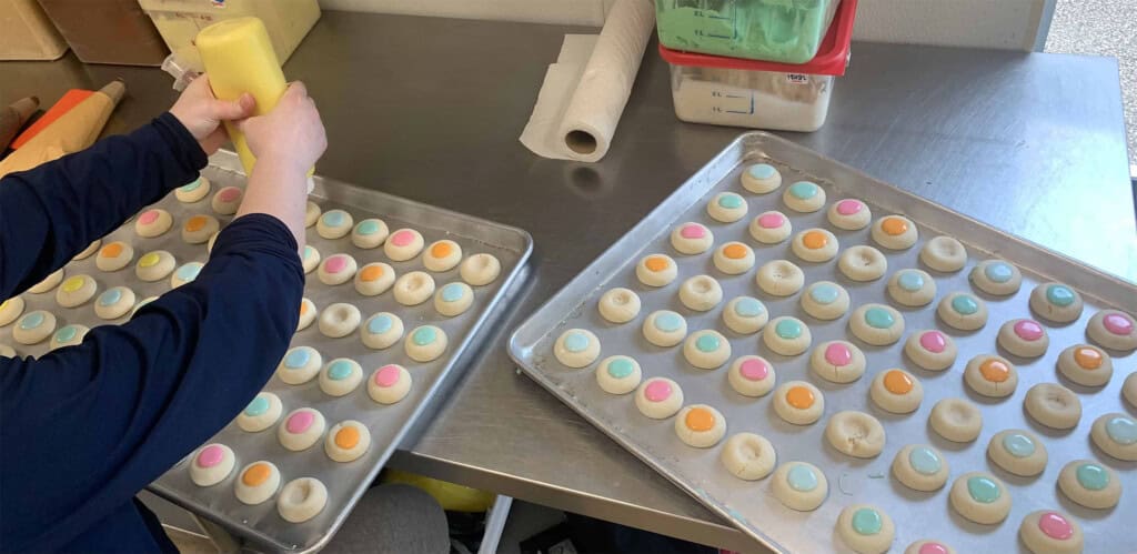 A person pipes yellow icing onto round cookies with colorful pastel centers. Two trays of cookies rest on a stainless steel counter in a bakery or kitchen setting.