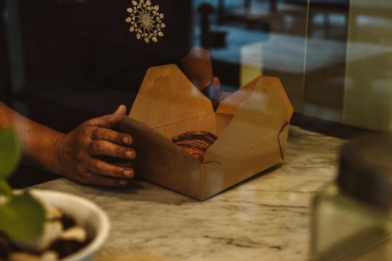 A person’s hand holds an open brown cardboard box with a pastry inside, resting on a marble countertop. The background is blurred, suggesting an indoor setting.