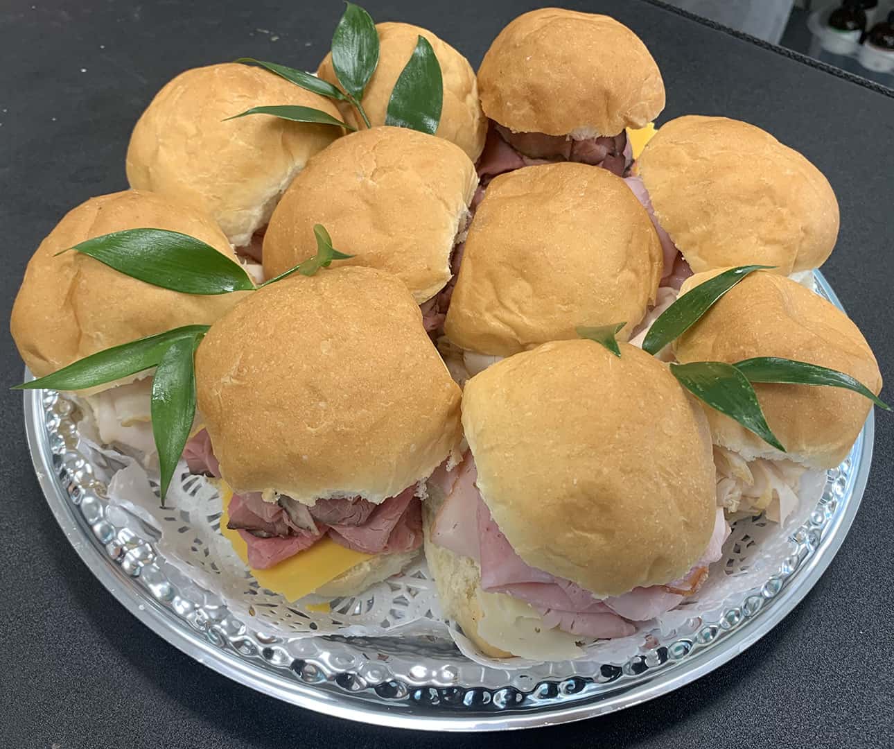 A platter of small sandwich rolls filled with assorted deli meats and cheeses, garnished with green leaves, arranged on a silver tray with a lace paper doily underneath.