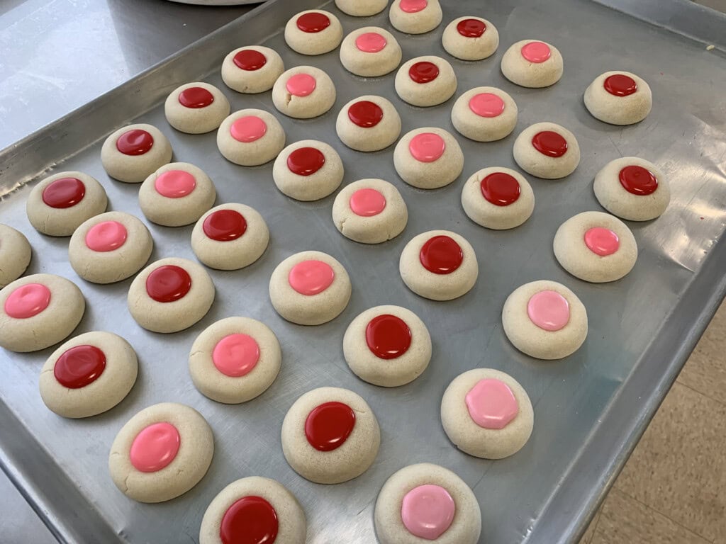 A baking tray filled with rows of thumbprint cookies. Each cookie has a colorful round center, some with pink filling and others with red filling. The cookies are evenly spaced on the metal tray.