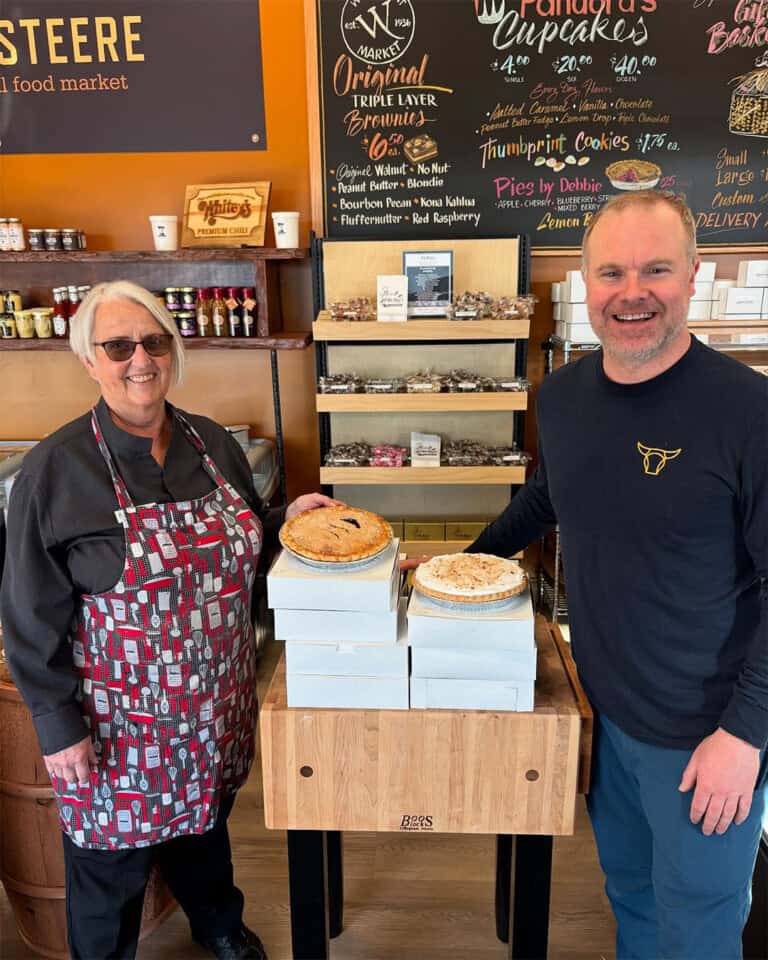 Two people stand smiling next to a table stacked with pie boxes and topped with two pies. They are inside a bakery or market, with shelves of baked goods and a chalkboard menu in the background.