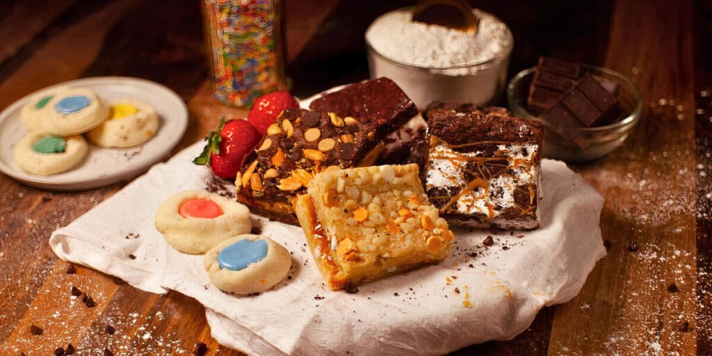An assortment of baked goods including brownies with toppings, a blondie bar with nuts, and colorful thumbprint cookies are arranged on a cloth, with baking ingredients like flour and chocolate nearby.