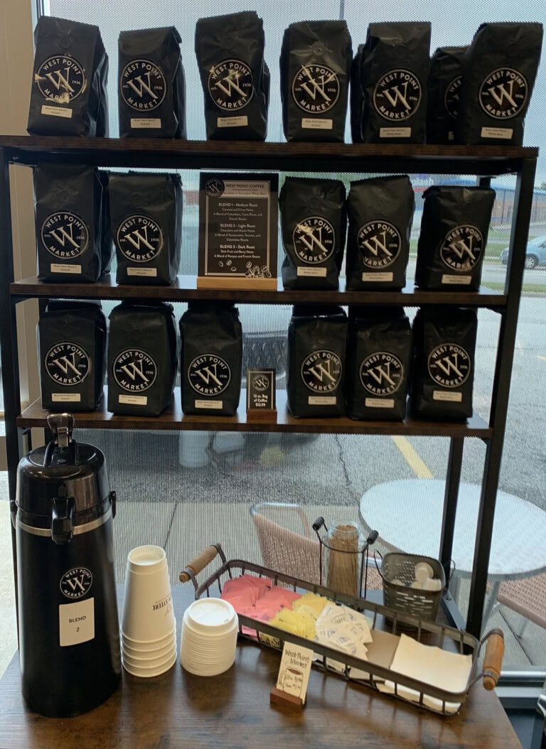 A coffee station with shelves holding multiple black bags of coffee, a large coffee dispenser, paper cups, lids, sweeteners, stirrers, napkins, and a small menu card, set near a window.