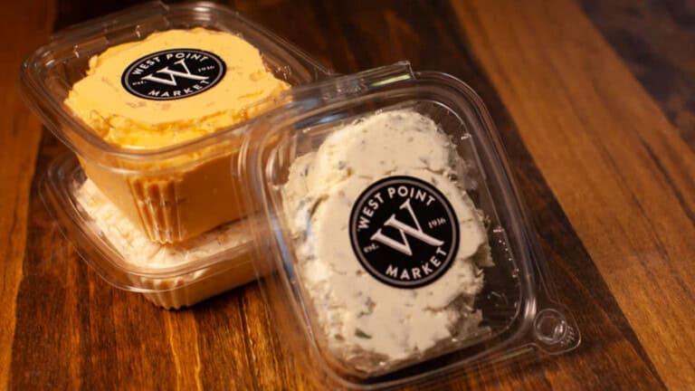 Two clear plastic containers with West Point Market labels, one filled with orange cheese spread and the other with white spread with herbs, stacked on a wooden surface.